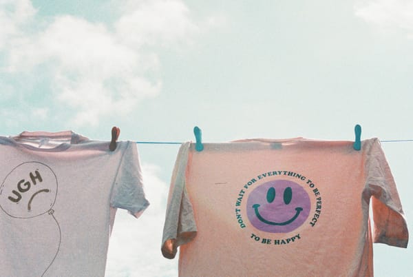 Two t-shirts drying. One saying UGH. Another with a happy smiley saying 'Don't want for everything to be perfect. Be happy'.